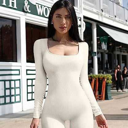 A woman with long dark hair wears the Miccelli Luxe Women's Seamless Long-Sleeve Yoga Jumpsuit in cream as she stands outdoors in front of a building with green and white trim.