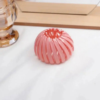 A pink Bird Nest Shaped Ponytail Hair Clip – Magic Lazy Braider Hairpin for Women by Miccelli Luxe rests on a white surface beside a gold-toned decor piece and a wooden-framed mirror.
