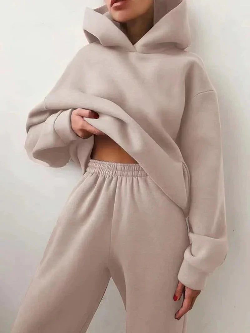 A model wears the Miccelli Luxe Two-Piece Tracksuit Set—a women’s light beige hooded jacket and matching sweatpants—posing against a plain white background and lifting the hoodie slightly to show part of her midriff.