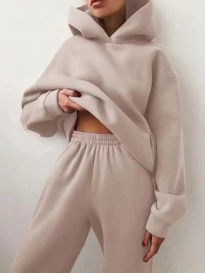 A model wears the Miccelli Luxe Two-Piece Tracksuit Set—a women’s light beige hooded jacket and matching sweatpants—posing against a plain white background and lifting the hoodie slightly to show part of her midriff.